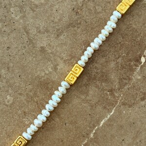 May include: A delicate necklace featuring small, white pearls strung together. The necklace is accented with rectangular gold-colored links with a Greek key design. The pearls are small and evenly spaced, creating a classic and elegant look.