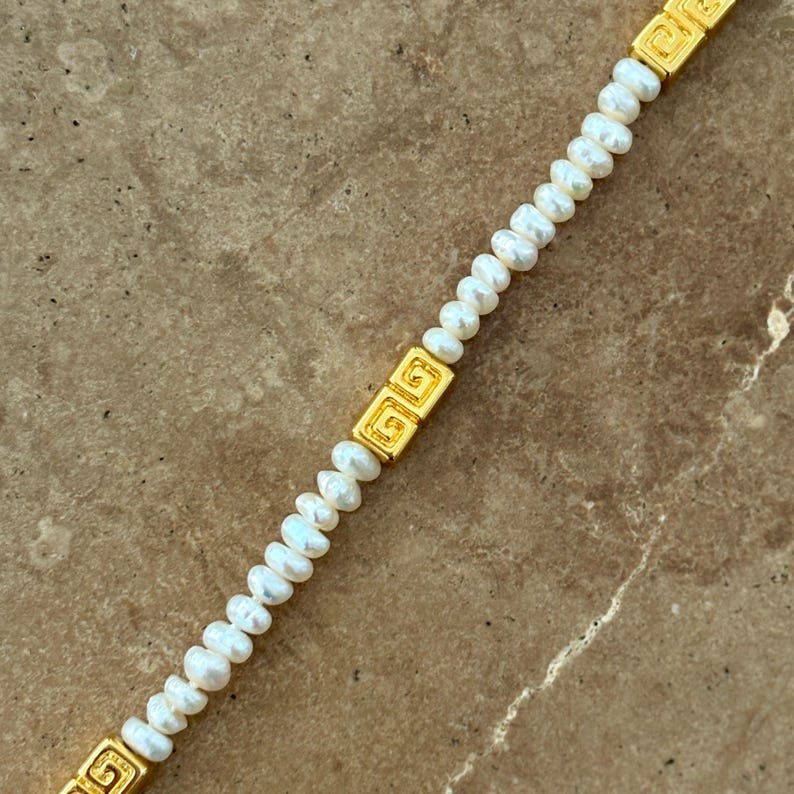 May include: A delicate necklace featuring small, white pearls strung together. The necklace is accented with rectangular gold-colored links with a Greek key design. The pearls are small and evenly spaced, creating a classic and elegant look.