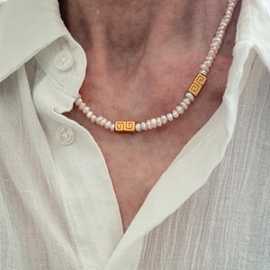 May include: A delicate pearl necklace with gold-colored square accents featuring a Greek key design. The necklace is worn against a white button-down shirt, creating a simple yet elegant look. The pearls are small and evenly spaced.