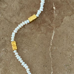 May include: A delicate necklace featuring small, white pearls strung together. The necklace is accented with two gold-colored rectangular clasps with a repeating square design. The jewelry is displayed on a brown surface.
