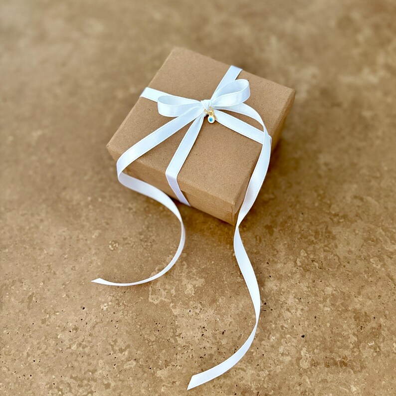 May include: A square brown cardboard gift box wrapped with a white satin ribbon tied in a bow. A small gold-toned charm featuring a blue and white evil eye symbol is attached to the center of the ribbon bow, resting on the top of the box.