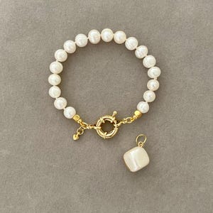 K&ouml;nnte beinhalten: Ein Armband aus wei&szlig;en Perlen mit einem goldenen Verschluss und einem quadratischen Perlenanh&auml;nger.