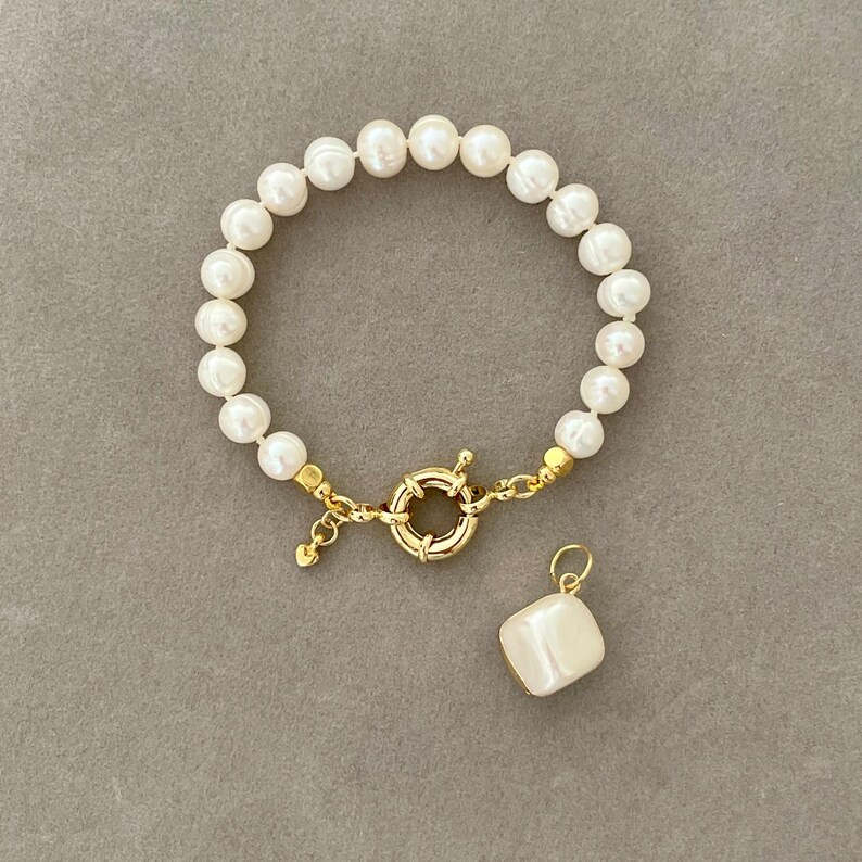 K&ouml;nnte beinhalten: Ein Armband aus wei&szlig;en Perlen mit einem goldenen Verschluss und einem quadratischen Perlenanh&auml;nger.