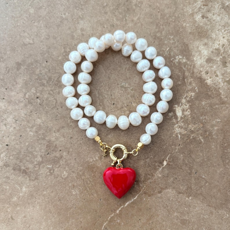 Peut inclure: Un collier compos&eacute; de perles d'eau douce blanches de forme irr&eacute;guli&egrave;re dispos&eacute;es en cercle. Le bijou pr&eacute;sente un fermoir circulaire dor&eacute; &agrave; l'avant, auquel est suspendu un pendentif en forme de c&oelig;ur rouge vif, lisse et brillant.