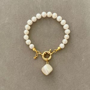K&ouml;nnte beinhalten: Ein Armband aus wei&szlig;en Perlen mit einem goldenen Verschluss und einem gro&szlig;en wei&szlig;en Perlenanh&auml;nger.