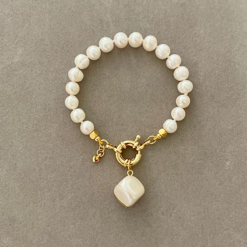 K&ouml;nnte beinhalten: Ein Armband aus wei&szlig;en Perlen mit einem goldenen Verschluss und einem gro&szlig;en wei&szlig;en Perlenanh&auml;nger.