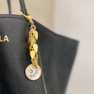Può includere: Ciondolo dorato con una moneta d'argento che presenta un disegno di un animale che corre. Il ciondolo è attaccato a una borsa tote in pelle nera con un design a foglia d'oro.