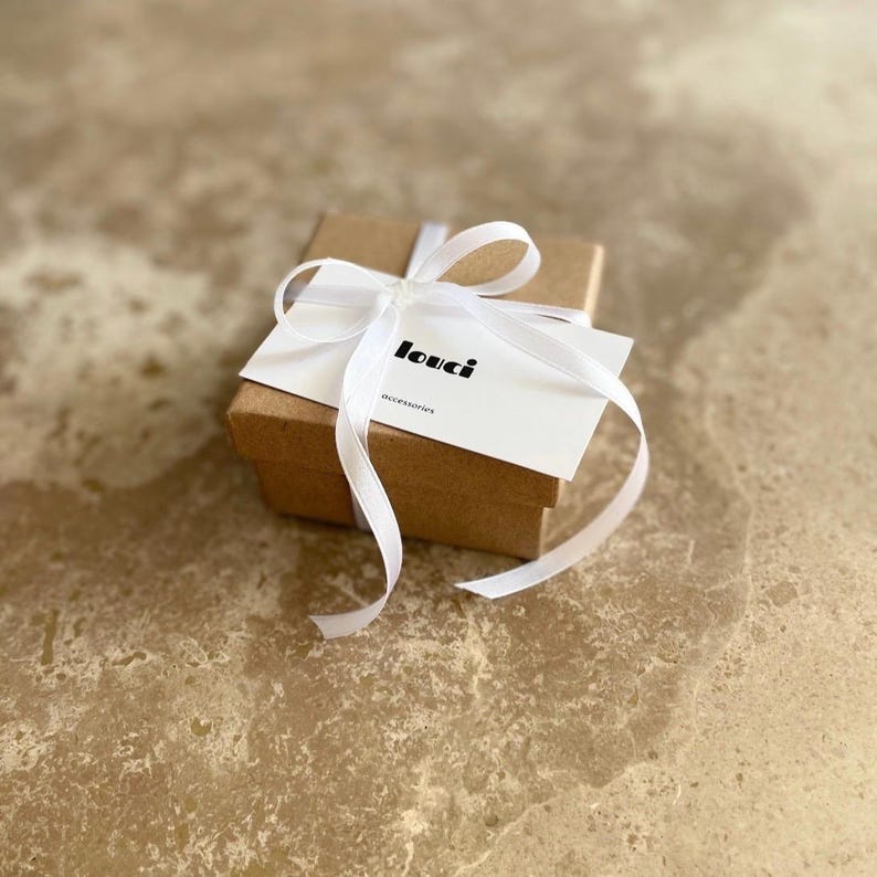 May include: A small, brown gift box tied with a white satin ribbon. A white card with the word "louci" and the word "accessories" is placed on top of the box. The box is on a light brown, textured surface.