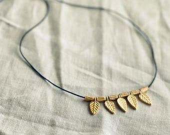 May include: A delicate necklace featuring five gold-colored leaf pendants strung on a thin, dark cord. The leaves are detailed with vein patterns, and the necklace has a simple, elegant design. The cord is adjustable.