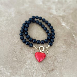 K&ouml;nnte beinhalten: Ein zweireihiges Armband mit glatten, runden schwarzen Perlen und einem goldfarbenen runden Verschluss. Ein einzelner, gl&auml;nzend roter herzf&ouml;rmiger Anh&auml;nger h&auml;ngt am Verschluss, erg&auml;nzt durch einen kleinen, zierlichen goldfarbenen Sternanh&auml;nger.