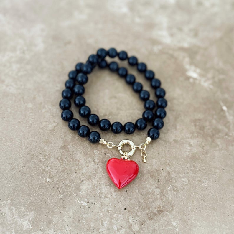 K&ouml;nnte beinhalten: Ein zweireihiges Armband mit glatten, runden schwarzen Perlen und einem goldfarbenen runden Verschluss. Ein einzelner, gl&auml;nzend roter herzf&ouml;rmiger Anh&auml;nger h&auml;ngt am Verschluss, erg&auml;nzt durch einen kleinen, zierlichen goldfarbenen Sternanh&auml;nger.