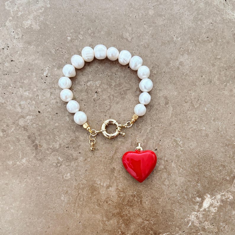 May include: A bracelet composed of white, round, lustrous pearls strung together, featuring a gold-toned circular clasp. A separate, smooth, glossy red heart-shaped charm with a gold-toned bail is positioned directly below the bracelet.