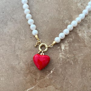 Peut inclure: Un collier compos&eacute; d'une rang&eacute;e de perles blanches sph&eacute;riques, d'un fermoir circulaire dor&eacute; et d'un pendentif en forme de c&oelig;ur rouge brillant. Une petite breloque &eacute;toile dor&eacute;e est fix&eacute;e au fermoir qui relie les deux extr&eacute;mit&eacute;s du collier.