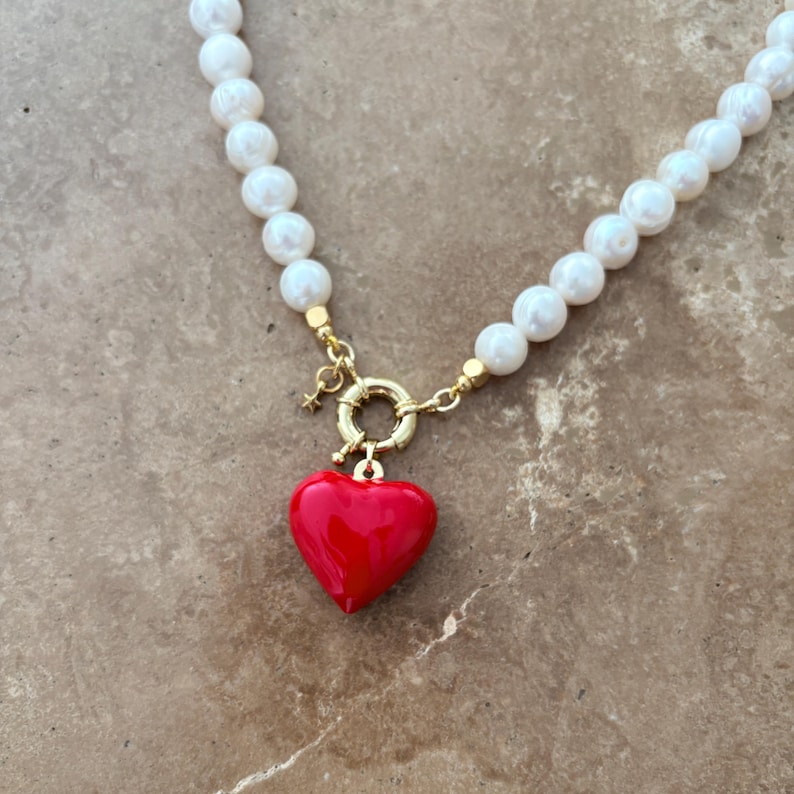 Peut inclure: Un collier compos&eacute; d'une rang&eacute;e de perles blanches sph&eacute;riques, d'un fermoir circulaire dor&eacute; et d'un pendentif en forme de c&oelig;ur rouge brillant. Une petite breloque &eacute;toile dor&eacute;e est fix&eacute;e au fermoir qui relie les deux extr&eacute;mit&eacute;s du collier.
