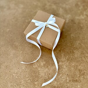 May include: A square brown cardboard gift box wrapped with a white satin ribbon tied in a bow. A small gold-toned charm featuring a blue and white evil eye symbol is attached to the center of the ribbon bow, resting on the top of the box.
