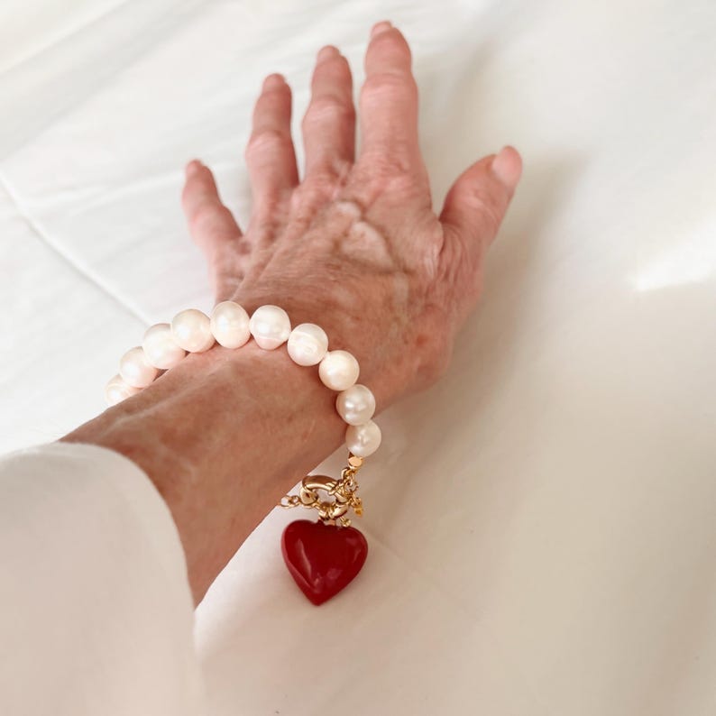 May include: A bracelet featuring a strand of round, off-white pearls with a gold-toned toggle clasp and a prominent, smooth, heart-shaped red charm hanging from the centre. The pearls are uniform in size and arranged in a circular pattern.