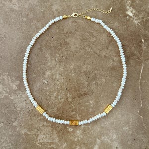 May include: A delicate necklace featuring small, white pearls strung together. The necklace is accented with three gold-colored, square-shaped decorative elements. The clasp and chain are also gold-colored, creating a cohesive and elegant design.
