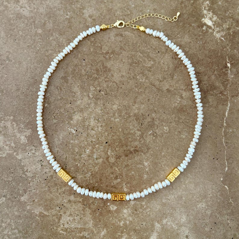 May include: A delicate necklace featuring small, white pearls strung together. The necklace is accented with three gold-colored, square-shaped decorative elements. The clasp and chain are also gold-colored, creating a cohesive and elegant design.