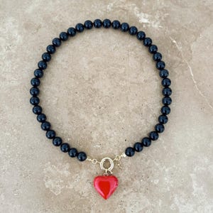 K&ouml;nnte beinhalten: Eine Perlenkette bestehend aus einem durchgehenden Strang glatter, runder, polierter schwarzer Perlen. Die Kette wird vorne mit einem goldfarbenen runden Verschluss geschlossen, an dem ein einzelner, gl&auml;nzender, hellroter herzf&ouml;rmiger Anh&auml;nger h&auml;ngt.
