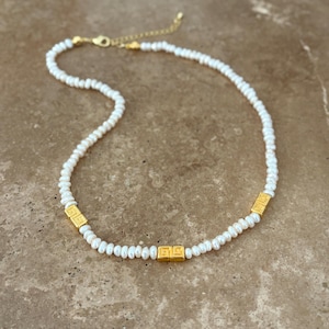 May include: A delicate necklace featuring small, white pearls strung together. The necklace is accented with three gold-colored square beads with a geometric design. The clasp and chain are also gold-colored, creating a classic and elegant look.