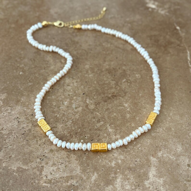 May include: A delicate necklace featuring small, white pearls strung together. The necklace is accented with three gold-colored square beads with a geometric design. The clasp and chain are also gold-colored, creating a classic and elegant look.