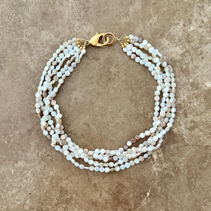 Puede incluir: Pulsera de múltiples hilos con pequeñas cuentas redondas de color blanco roto. La pulsera tiene un cierre y herrajes de conexión dorados. Las cuentas tienen sutiles variaciones de color, con toques de rosa y marrón.
