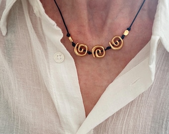 May include: A necklace featuring three gold-toned spiral charms strung on a thin black cord. The spiral elements are evenly spaced along the cord, accented by small gold-toned beads on either side of the central arrangement, creating a simple design.