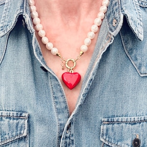 Peut inclure: Un collier compos&eacute; d'un rang de perles blanches, rondes et lustr&eacute;es, reli&eacute; &agrave; un fermoir circulaire dor&eacute;. Un pendentif en forme de c&oelig;ur rouge vif, lisse et brillant, est suspendu au fermoir, accompagn&eacute; d'une petite breloque &eacute;toile dor&eacute;e.