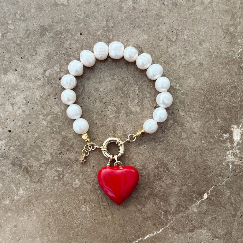 May include: A bracelet composed of white, irregularly shaped freshwater pearls strung together, featuring a gold-toned circular clasp and a prominent, smooth, glossy red heart-shaped charm hanging from the centre of the clasp mechanism.