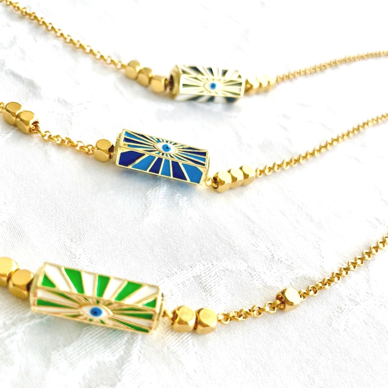 May include: Three gold necklaces with a chain and a rectangular pendant with a blue evil eye design. The pendants are white with blue, green, and white stripes.