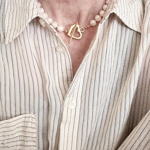May include: A necklace featuring a strand of white pearls and a gold-toned heart-shaped toggle clasp, worn with a cream-colored button-down shirt that has thin, vertical black pinstripes and visible white buttons along the front placket.