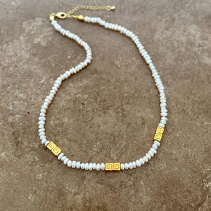May include: A delicate necklace featuring small, white pearls strung together. The necklace is accented with three gold-colored rectangular beads, each with a repeating geometric design. The clasp and chain are also gold-colored.