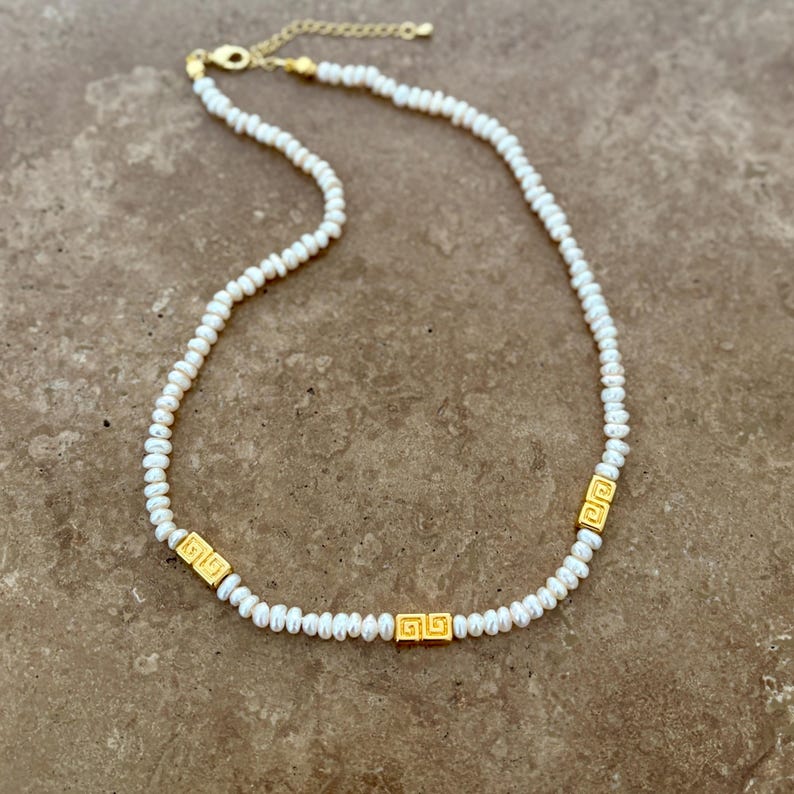 May include: A delicate necklace featuring small, white pearls strung together. The necklace is accented with three gold-colored rectangular beads, each with a repeating geometric design. The clasp and chain are also gold-colored.