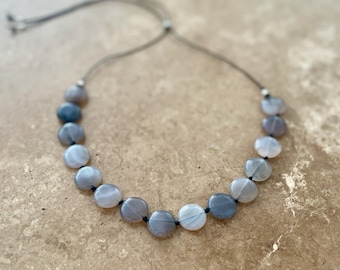 May include: A gray and black gemstone necklace with a black cord. The necklace is made of round, flat stones that are graduated in size. The stones have a subtle shimmer.