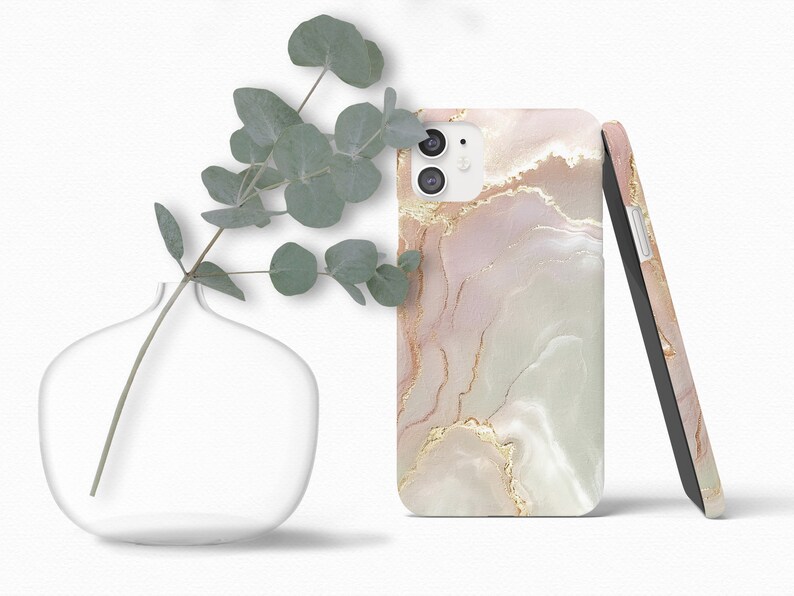 May include: A pair of phone cases with a marble design in shades of pink, gold, and white. One case is upright, and the other is leaning against it. A clear glass vase with eucalyptus branches is in the background.