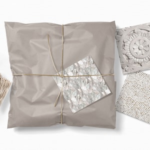 Puede incluir: Un paquete de papel marr&oacute;n atado con cuerda dorada. El paquete est&aacute; decorado con tres peque&ntilde;as muestras cuadradas de azulejos decorativos. Un azulejo es un mosaico blanco y gris, otro es un patr&oacute;n de cuentas blancas y doradas, y el &uacute;ltimo es un dise&ntilde;o floral blanco tallado.
