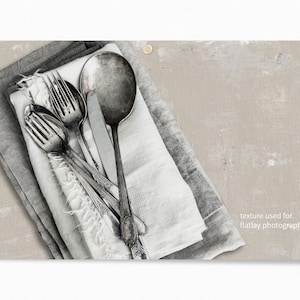 May include: A close-up of a set of silver cutlery, including a fork, knife, and spoon, arranged on a white and gray linen napkin. The cutlery is arranged in a criss-cross pattern, with the knife placed on top of the fork and spoon. The cutlery is slightly tarnished, giving it a vintage look. The background is a light gray, textured surface.  texture used for flatlay photography