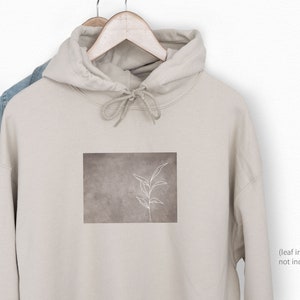 Op de afbeelding: Een beige hoodie met een witte omtrek van een blad op een bruine achtergrond. De hoodie heeft een capuchon met een trekkoord.