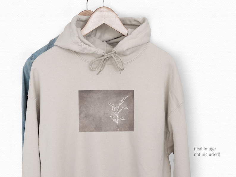 Op de afbeelding: Een beige hoodie met een witte omtrek van een blad op een bruine achtergrond. De hoodie heeft een capuchon met een trekkoord.