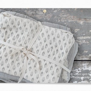 Op de afbeelding: Een stapel witte stof met een zwart bloemenpatroon is vastgebonden met een wit lint. De stof ligt op een grijs houten oppervlak. "texture used for flatlay photography" staat in zwarte tekst rechtsonder in de afbeelding.