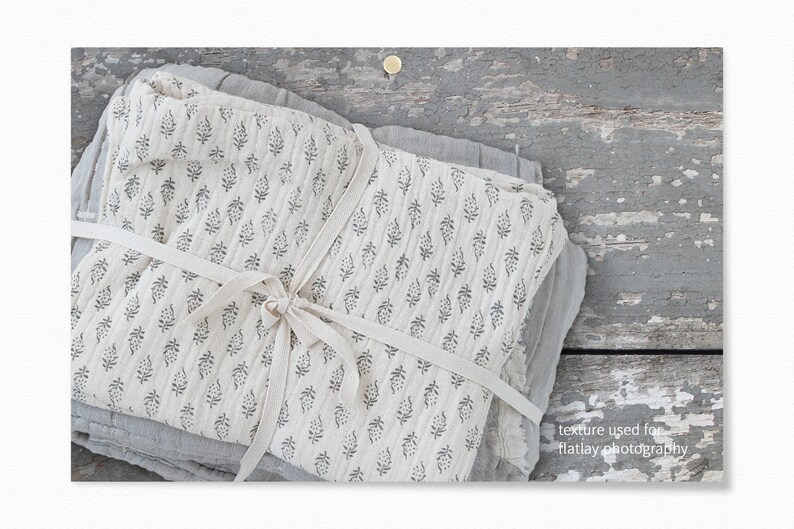 Op de afbeelding: Een stapel witte stof met een zwart bloemenpatroon is vastgebonden met een wit lint. De stof ligt op een grijs houten oppervlak. "texture used for flatlay photography" staat in zwarte tekst rechtsonder in de afbeelding.