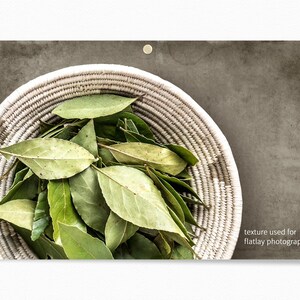 May include: A white woven basket filled with fresh green bay leaves. The basket is on a textured gray background.