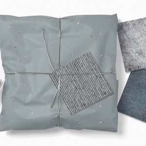 Op de afbeelding: Een grijs cadeau ingepakt in zilverpapier met zilveren sterren en vastgebonden met zilveren touw. Het cadeau ligt op een wit oppervlak. Er zijn drie kleine vierkanten stof op de achtergrond, een is grijs, een is blauw en een is lichtgrijs met een geweven textuur.