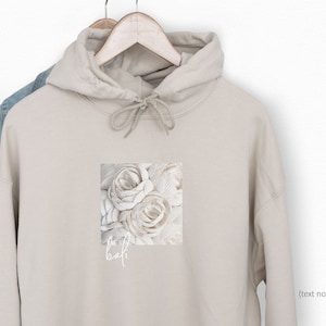 Puede incluir: Una sudadera con capucha beige con un dise&ntilde;o floral blanco y el texto "the half" impreso en la parte delantera.