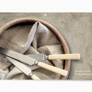 Op de afbeelding: Drie zilveren botermesjes met ivoor handvatten zijn gerangschikt in een houten kom bekleed met een beige linnen doek. De kom staat op een getextureerd grijs oppervlak. "textuur gebruikt voor flatlay fotografie" staat in witte tekst in de rechterbenedenhoek van de afbeelding.