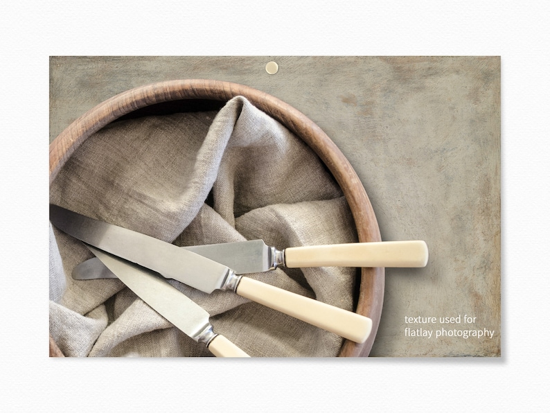 Op de afbeelding: Drie zilveren botermesjes met ivoor handvatten zijn gerangschikt in een houten kom bekleed met een beige linnen doek. De kom staat op een getextureerd grijs oppervlak. "textuur gebruikt voor flatlay fotografie" staat in witte tekst in de rechterbenedenhoek van de afbeelding.