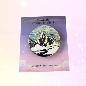 May include: A round, gold-rimmed e-book grip featuring a mountain range design with a blue and gold color scheme. The text "Bookish E-BOOK GRIP" is at the top. The website address www.lovinbookscandle.de is at the bottom.