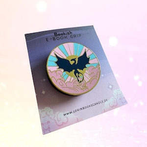 May include: A round, black E-BOOK GRIP with a dragon design. The dragon is black, with a gold sun and pink and blue clouds in the background. The text "Bookish E-BOOK GRIP" is at the top.