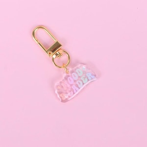 Könnte beinhalten: Ein klarer Acryl-Schlüsselanhänger mit den Worten "HOOD READER" in Pastellfarben. Der Schlüsselanhänger hat einen goldfarbenen Verschluss und Ring, vor einem rosa Hintergrund.