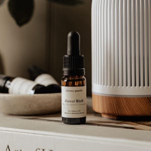 May include: A white, ribbed, dome-shaped electric diffuser with a wood-grain base sits next to a small amber glass dropper bottle labelled 'aroma quartz Forest Walk'. The bottle features a black dropper cap and sits on a book titled The Art of Home.